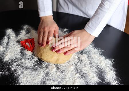 Woman kneads cake dough with hands on the table sprinkled with flour and a red mold in the shape of Christmas tree on the desk at home Stock Photo