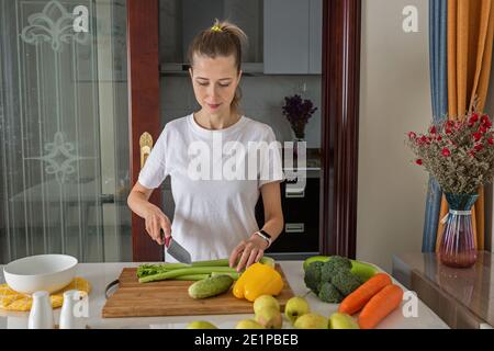 Young caucasian woman cooking fresh fruit and vegetable salad on table. Person preparing healthy yummy eating lunch in kitchen during coronavirus Stock Photo