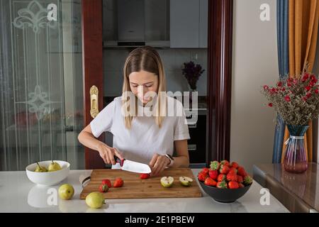 Young caucasian woman cooking fresh fruit and vegetable salad on table. Person preparing healthy yummy eating lunch in kitchen during coronavirus Stock Photo