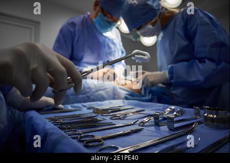 Focus on doctor's hand holding forceps with tampon while surgeon and assistant performing plastic surgery. Medical team doing aesthetic surgery and using various surgical tools in operating room. Stock Photo