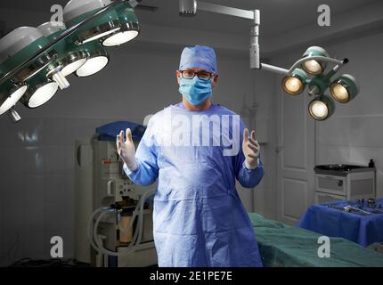 Portrait of surgeon looking at camera while standing in operating room at hospital. Man doctor wearing surgical uniform, sterile gloves, protective face mask, medical cap. Concept of medicine, surgery Stock Photo