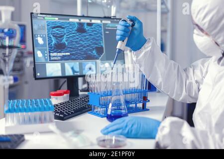 Medic assistant working in biochemistry lab working with automatic pipette wearing protection suit. Chemist in modern laboratory doing research using dispenser during global epidemic with covid-19. Stock Photo