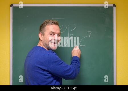 Happy smiling male teacher giving a mathematics class turning to look at his students after writing an equation on the blackboard Stock Photo