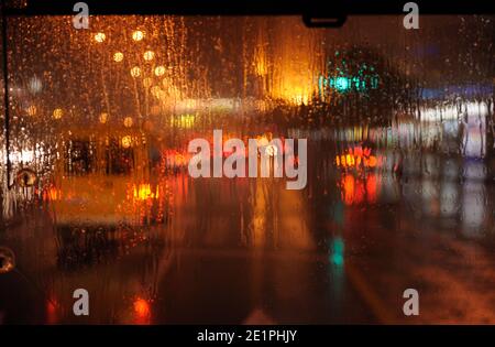 Drops and trickles of rain water dripping on car's windscreen at night. Blurred street traffic with warm light in background. Stock Photo