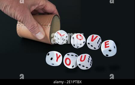 White dice with black eyes numbers spelling LOVE YOU  in red letters rolling out from a raffle cup held by a hand isolated on black background. Love a Stock Photo