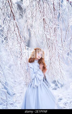 Young redhead woman, a princess, walks in a winter forest in a blue dress. Stock Photo