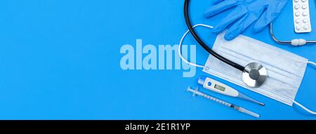 Stethoscope, face mask, medical syringe, thermometer, pills blister and medical gloves on a blue background. Flat lay banner with copy space. Stock Photo