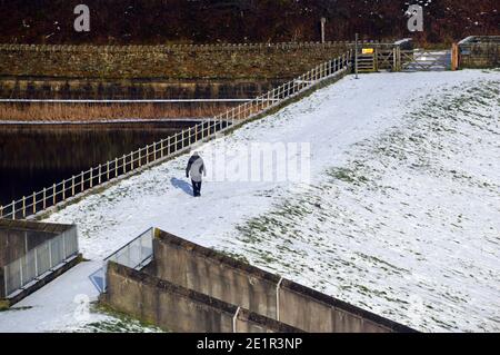 Man Walking in Snow on Lower Ogden Reservoir Dam near the Village of Barley from Path to Pendle Hill in Ogden Clough, Lancashire. UK. Stock Photo