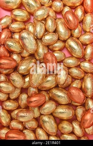 Large and small pile or group of gold, orange and bronze foil wrapped chocolate easter eggs, against a peach orange background. Stock Photo