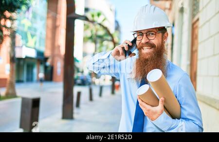 Young redhead architect man with long beard wearing hardhat smiling happy. Having conversation talking on the smartphone holding blueprints at street Stock Photo