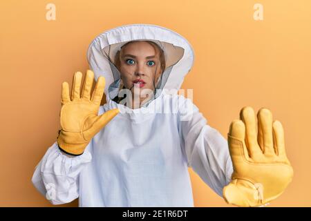 Beautiful blonde caucasian woman wearing protective beekeeper uniform afraid and terrified with fear expression stop gesture with hands, shouting in s Stock Photo
