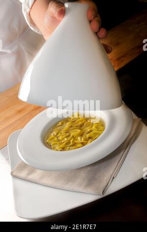 Ravioli del plin in broth, typical Piedmontese stuffed pasta from the Langhe, chef hand lift up the ceramic bell on the hot dish Stock Photo