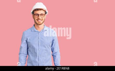 Handsome young man with bear wearing architect hardhat looking positive and happy standing and smiling with a confident smile showing teeth Stock Photo