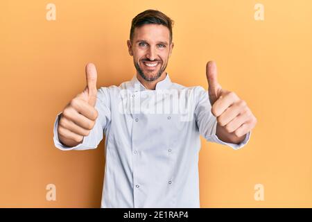 Handsome man with beard wearing professional cook uniform approving doing positive gesture with hand, thumbs up smiling and happy for success. winner Stock Photo