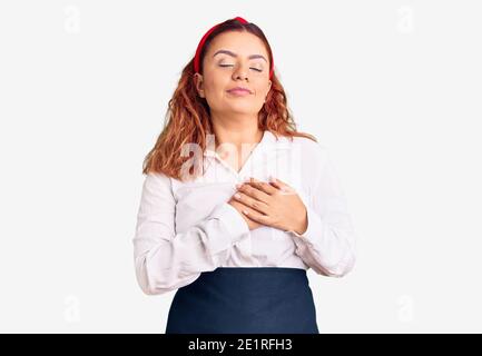 Young latin woman wearing waitress apron smiling with hands on chest with closed eyes and grateful gesture on face. health concept. Stock Photo