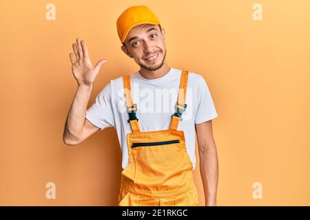 Hispanic young man wearing handyman uniform waiving saying hello happy and smiling, friendly welcome gesture Stock Photo