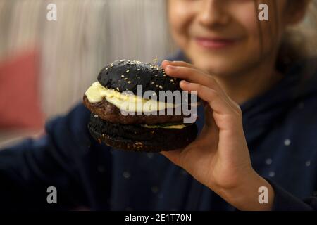 Black cheeseburger in the hands of a girl close-up in a cafe. The concept of children's food Stock Photo