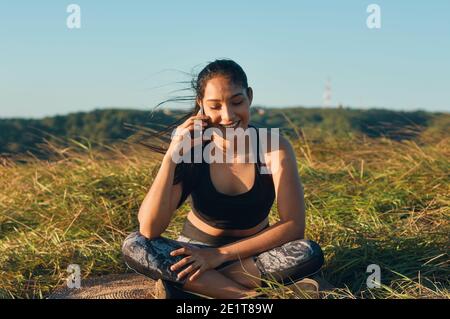 A happy young woman sitting on the grass and talking on the phone after workout Stock Photo