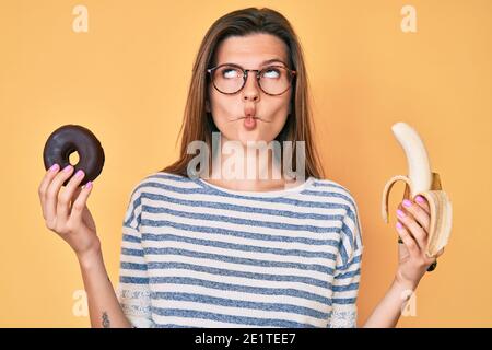 Beautiful caucasian woman holding banana and chocolate doughnut making fish face with mouth and squinting eyes, crazy and comical. Stock Photo