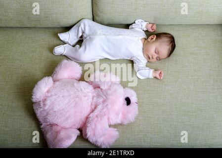 Baby sleeps in room, infant dreams on couch at home. Top view of sleeping small baby and pink soft toy on sofa. Little child in romper lying on his ba Stock Photo