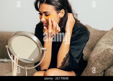 Young woman in sating robe sitting on a couch in front of a mirror applying an eye patch Stock Photo