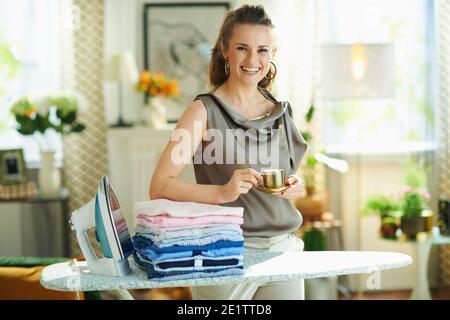 Portrait of happy middle age woman in silk blouse and beige pants with steam iron, ironing board and pile of folded ironed clothes drinking cup of esp Stock Photo