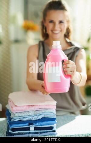 Smiling modern woman in silk blouse and beige pants with ironing board, pile of folded ironed clothes and pink bottle of fabric conditioner in the mod Stock Photo