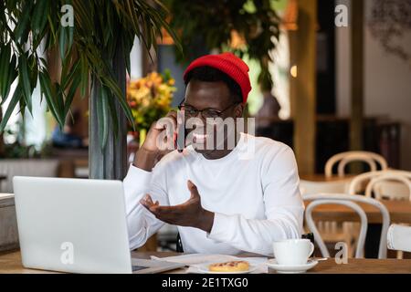 Happy millennial Black man laughing, speaking with friend on mobile phone, remote work in cafe. Stock Photo