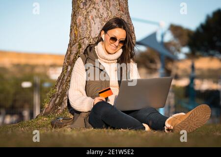 Happy woman sitting outdoors holding credit card and using laptop computer. Online shopping Stock Photo