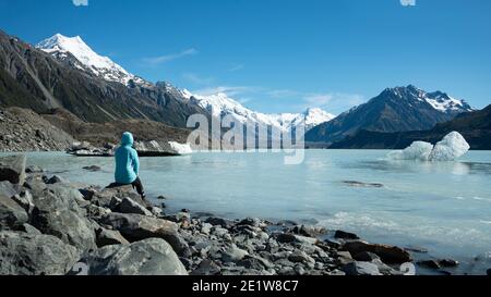 Tourist sitting on the shore of Tasman Glacier terminal lake looking at snow-capped mountains and icebergs Stock Photo