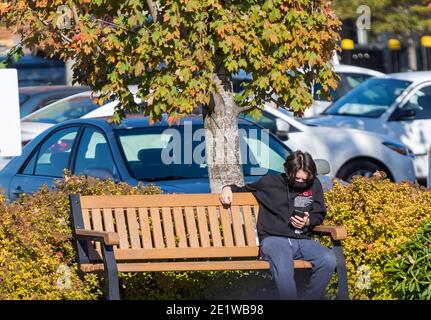 Young man with a mask sitting on a bench in a city typing messages with his phone. White Rock, British Columbia/Canada-October 22,2020. Street photo, Stock Photo
