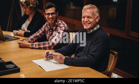 Senior businessman in boardroom meeting with colleagues sitting by. Happy male professional sitting at conference table and smiling. Stock Photo