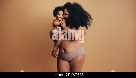 Plus size woman with her baby. Happy mommy carrying her toddler in studio. Real woman body after child birth. Stock Photo