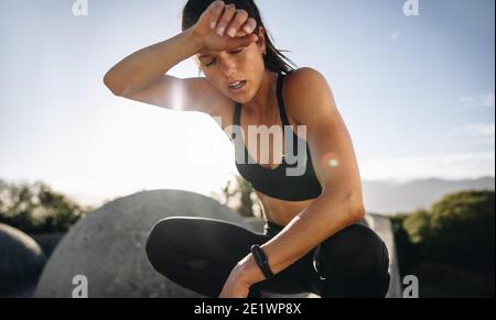 Tired woman sitting and resting after workout. Woman feeling exhausted after training session wiping her sweat from her forehead. Stock Photo