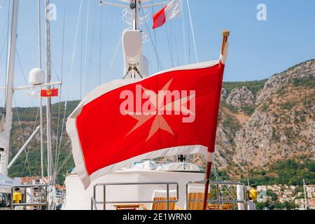 The Maltese flag on a flagpole flies in the wind on board a yacht against the backdrop of mountains. Stock Photo