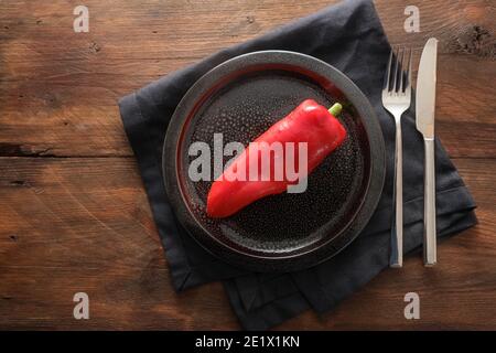Raw red pointed sweet pepper on a dark plate, cutlery and napkin on a rustic wooden background, healthy eating concept or vegetable diet to lose weigh Stock Photo