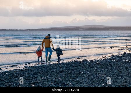 Rear view of father holding hands with his two young children walking along coast during winter, Lepe, Hampshire, England Stock Photo