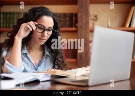 Young lady reading book sitting at desk touching glasses Stock Photo
