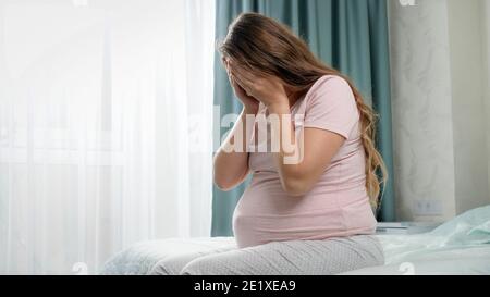 Sad crying pregnant woman suffering from depression sitting on bed and holding her head. Concept of maternal and pregnancy depression. Stock Photo