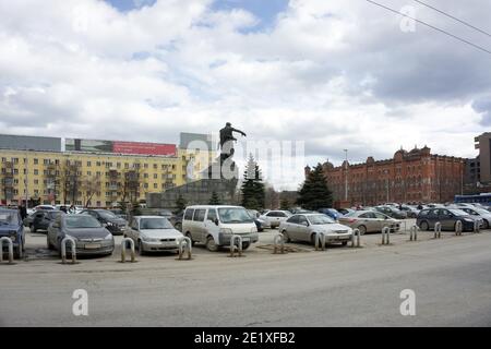Railway station square with a monument to the soldiers of the Ural Volunteer Tank Corps (1962) and parked cars nearby on a cloudy spring day.