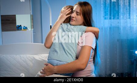 Young loving pregnant mother hugging and talking to her sad and scared toddler son lying in bed and trying to sleep at night. Parent soothing child Stock Photo