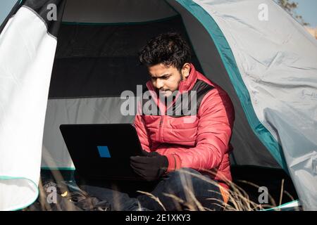 Young man busy working on laptop inside the tent during camping - concept of digital nomadic lifestyle and solo travel. Stock Photo