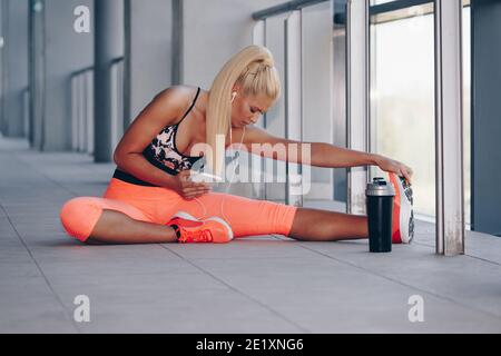 Determinate girl doing stretching in building. Woman in fitness gear using smartphone and  listening to music using earphones. Stock Photo