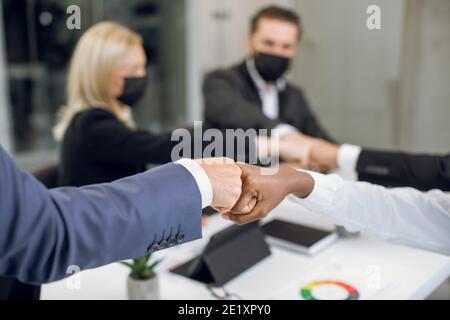 Close up cropped image of happy business coworkers with face protective masks, bumping their fists after successful meeting in the office. Focus on Stock Photo