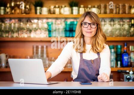 Confident mature small cafe owner businesswoman standing behind the counter and using laptop while working. Stock Photo