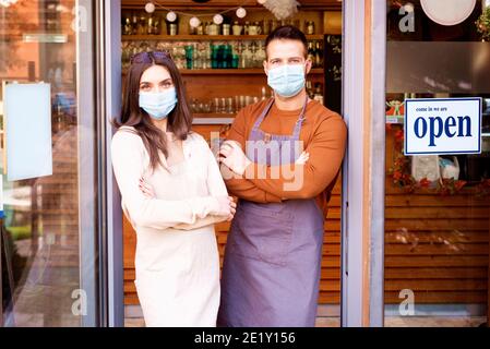 Shot of young waitress and waiter wearing face mask for prevention while standing in the restaurant’s doorway after reopening coronavirus pandemic. Op Stock Photo