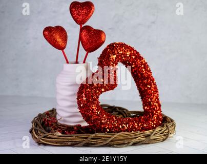 Valentine’s Day Decor, wooden heart wreath, red heart wreath, white vase filled with red glittery hearts sticking up on a white and gray background. Stock Photo