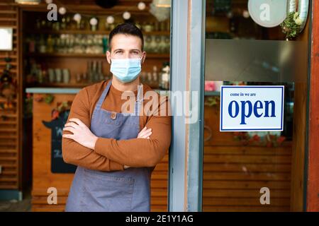 Portrait of smiling owner man wearing face mask for prevention while standing at his cafe door with open signboard. Stock Photo