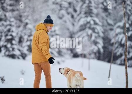 Cheerful young man with dog in winter. Pet owner with his labrador retriever walking in snow against forest. Jizera mountains, Czech Republic Stock Photo