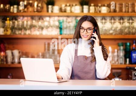 Shot of happy female coffee shop owner standing in the coffee shop and working. Young waitress using mobile phone and laptop. Stock Photo